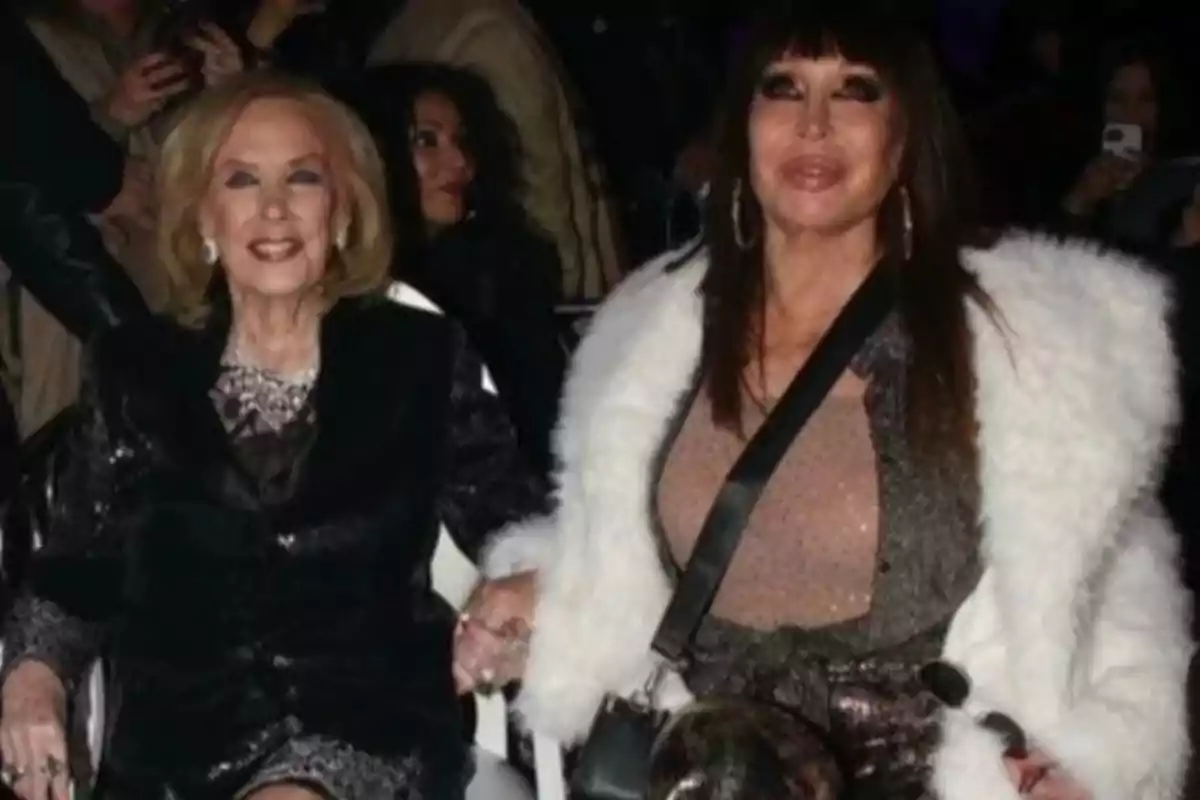 Two elegantly dressed women are sitting together at an event, holding hands and smiling, surrounded by other people in the background. Two elegantly dressed women are sitting together at an event, holding hands and smiling, surrounded by other people in the background.