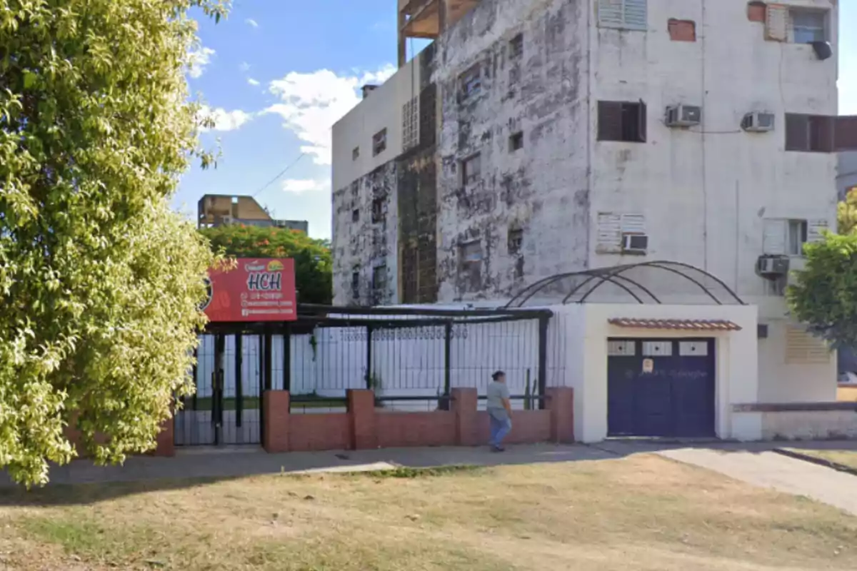 Facade of a multi-story building with worn walls, a black gate, a red sign for a store, and a person walking on the sidewalk on a sunny day. Facade of a multi-story building with worn walls, a black gate, a red sign for a store, and a person walking on the sidewalk on a sunny day.