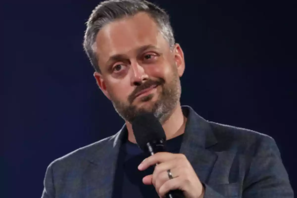 Man with a beard and short hair holds a microphone while looking ahead with a thoughtful expression. Man with a beard and short hair holds a microphone while looking ahead with a thoughtful expression.