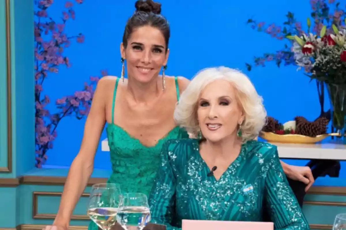 Two elegantly dressed women in green pose smiling on a television set decorated with flowers and wine glasses on the table.