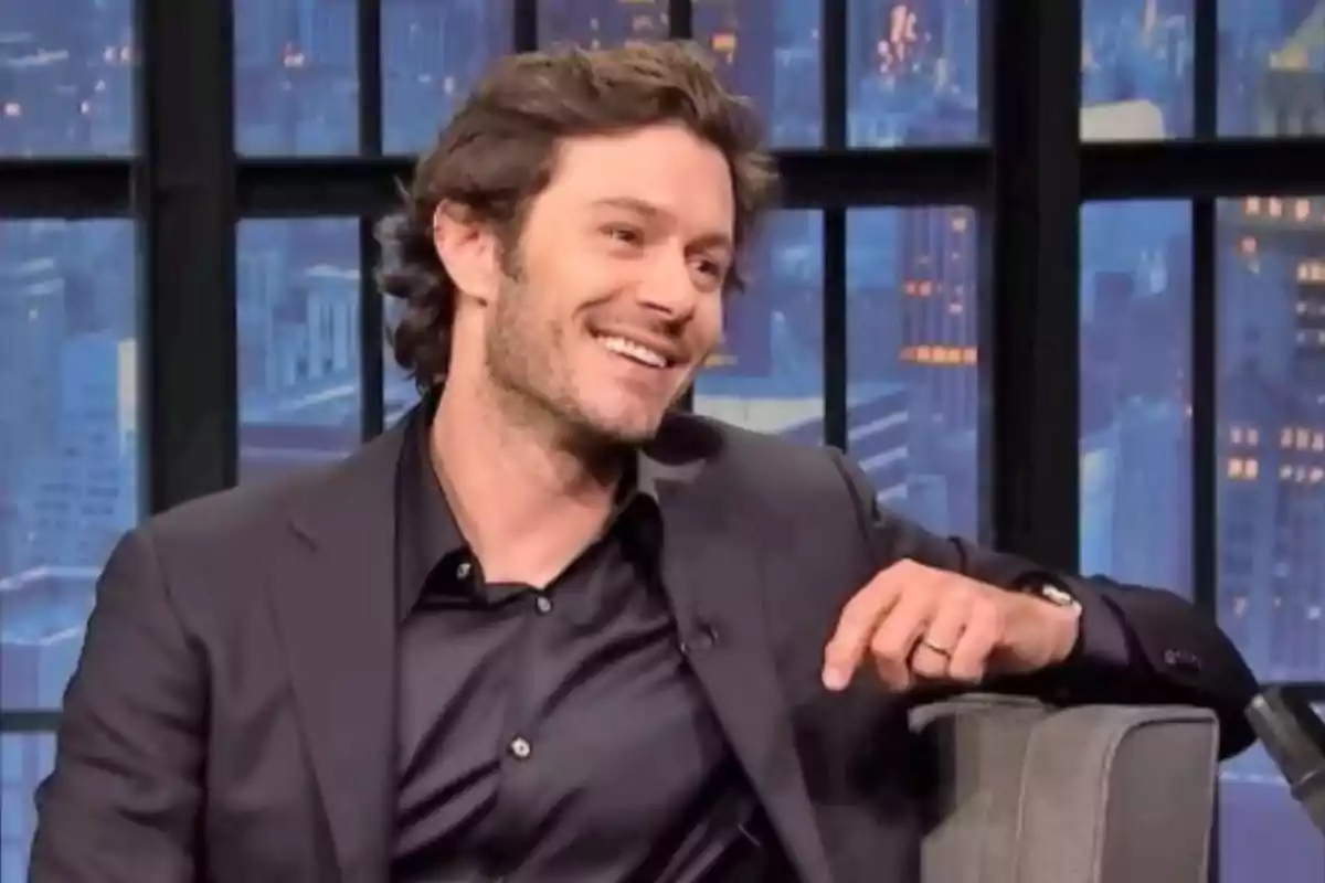 Young man with brown hair and a light beard smiling while sitting on a television set with windows and buildings in the background Young man with brown hair and a light beard smiling while sitting on a television set with windows and buildings in the background
