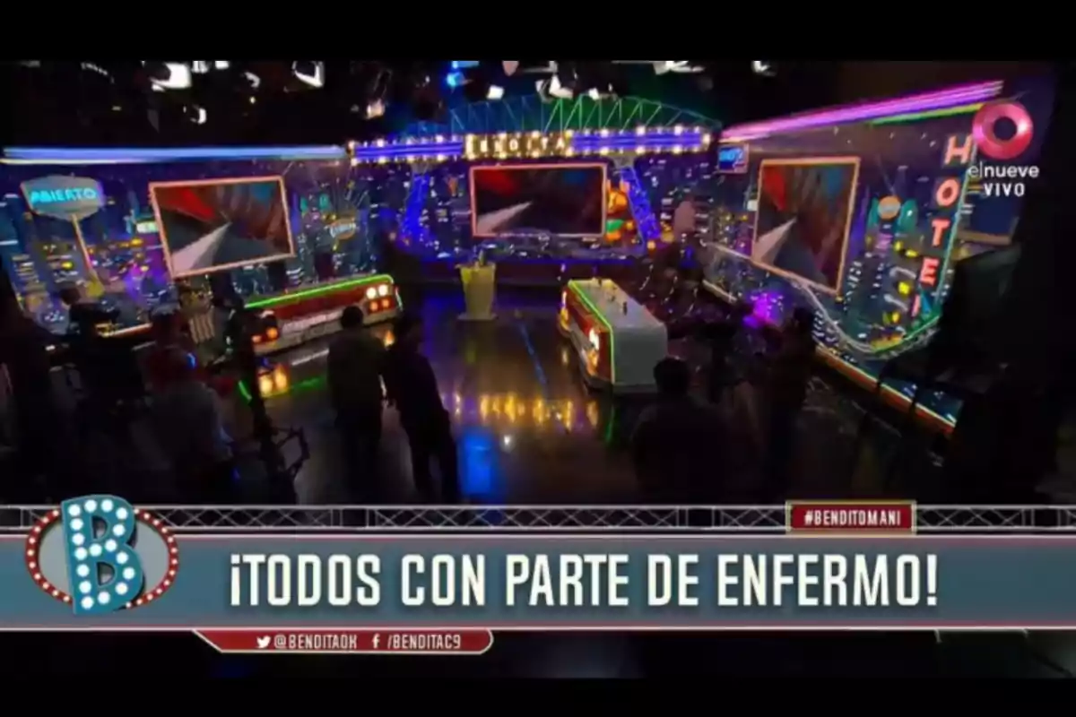 Colorful TV set with several screens and lights on, people in the studio, and a big sign that says “Everyone with a sick note!” Colorful TV set with several screens and lights on, people in the studio, and a big sign that says “Everyone with a sick note!”
