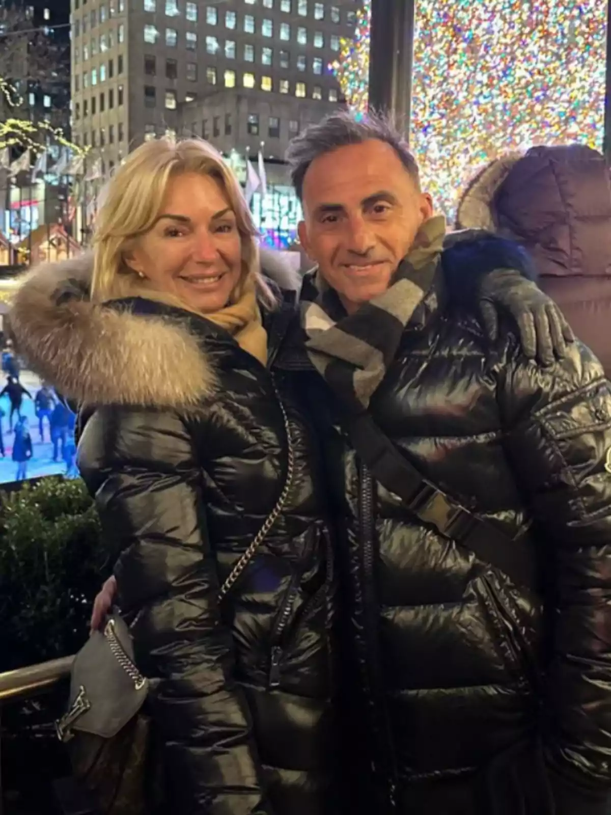 Bundled-up couple smiling while posing in front of a lit Christmas tree in a city at night Bundled-up couple smiling while posing in front of a lit Christmas tree in a city at night