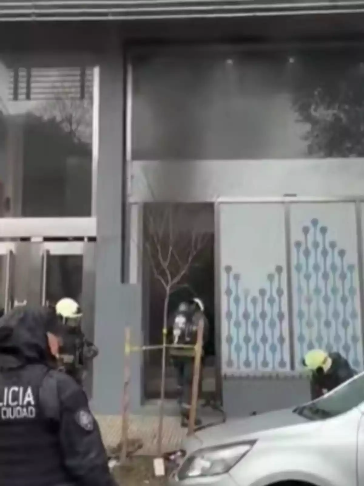 Several firefighters and police officers are responding to an emergency at the entrance of a building while smoke comes out from inside and yellow tape restricts access. Several firefighters and police officers are responding to an emergency at the entrance of a building while smoke comes out from inside and yellow tape restricts access.