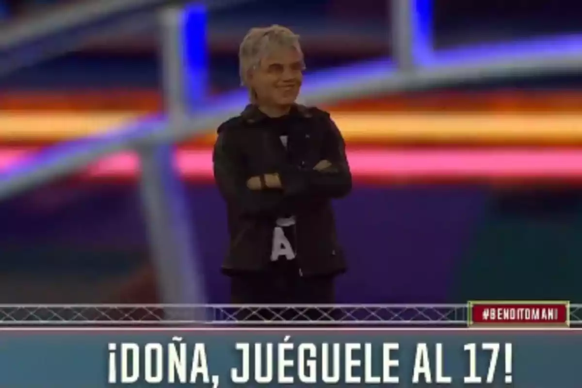 Smiling person with crossed arms on a colorful set and text at the bottom that says Doña, play 17 Smiling person with crossed arms on a colorful set and text at the bottom that says Doña, play 17