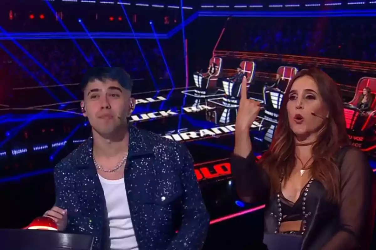 Dos jueces de un programa de talentos están sentados en sus sillas, uno de ellos presiona un botón rojo mientras la otra persona hace un gesto con la mano y parece estar hablando, el escenario está iluminado con luces azules y rojas