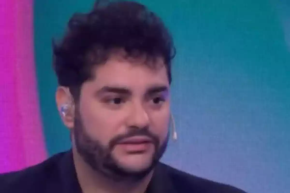 Hombre de cabello oscuro y barba usando un micrófono de diadema en un set de televisión con fondo colorido
