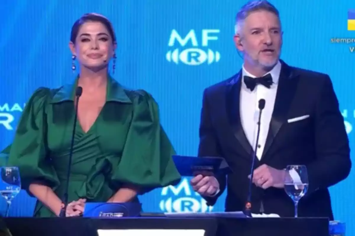 Dos presentadores elegantemente vestidos están en un escenario frente a micrófonos y copas de agua, con un fondo azul y logotipos detrás de ellos.