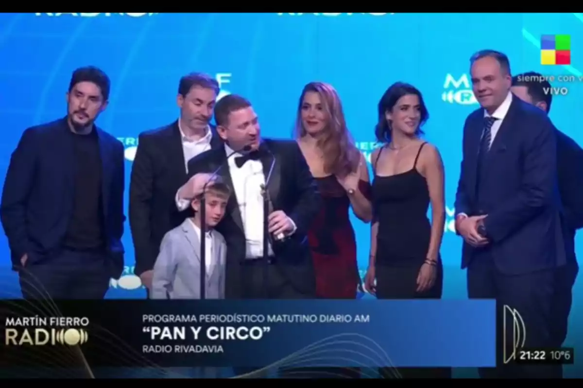 Un grupo de personas elegantemente vestidas celebra en el escenario durante una premiación, con fondo azul y el texto Pan y Circo Radio Rivadavia en la pantalla. Un grupo de personas elegantemente vestidas celebra en el escenario durante una premiación, con fondo azul y el texto Pan y Circo Radio Rivadavia en la pantalla.