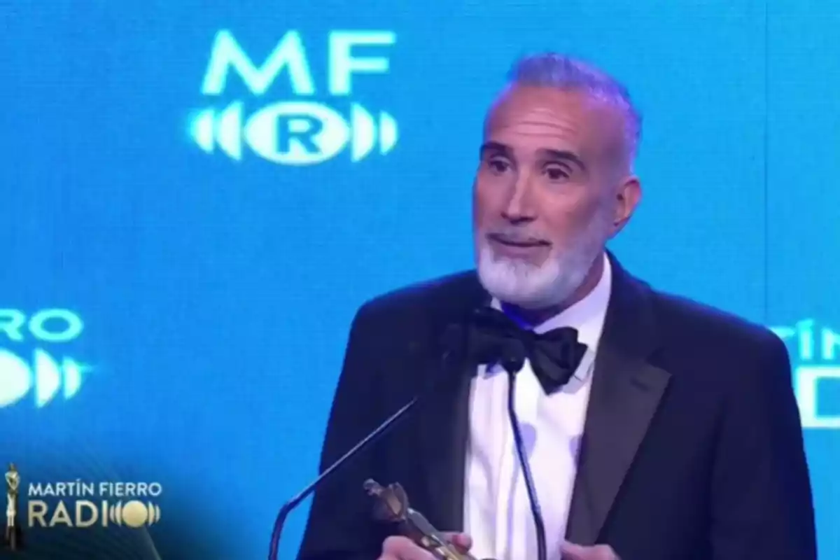 Hombre de barba canosa con esmoquin sosteniendo un premio y hablando frente a un micrófono en una ceremonia con fondo azul
