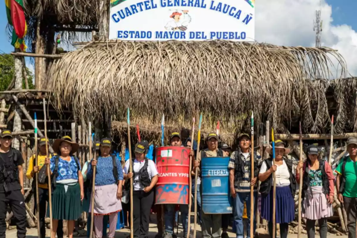 A group of people with spears and shields made from metal barrels pose in front of a wooden structure with a palm roof and a sign that reads Lauca Ñ Headquarters General Staff of the People. A group of people with spears and shields made from metal barrels pose in front of a wooden structure with a palm roof and a sign that reads Lauca Ñ Headquarters General Staff of the People.