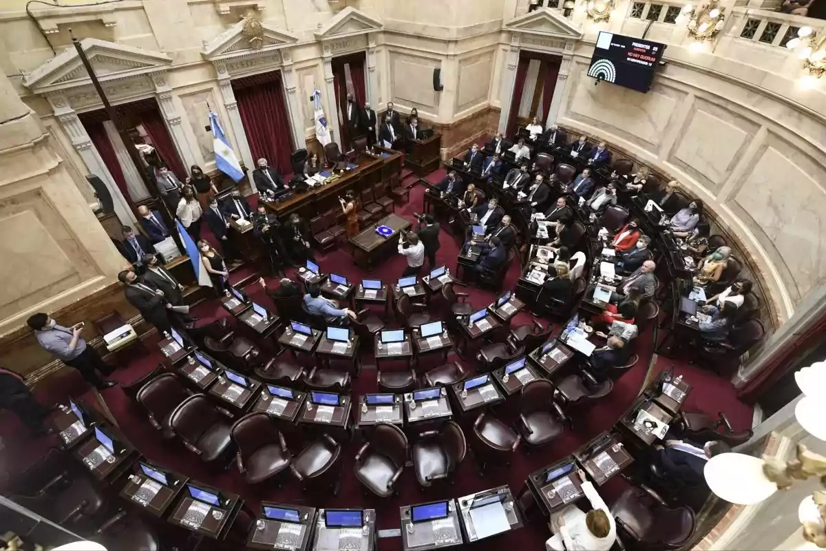 Vista general del recinto del Senado argentino durante una sesión con varios legisladores sentados y otros de pie, rodeados de escritorios y pantallas en un ambiente formal y solemne Vista general del recinto del Senado argentino durante una sesión con varios legisladores sentados y otros de pie, rodeados de escritorios y pantallas en un ambiente formal y solemne