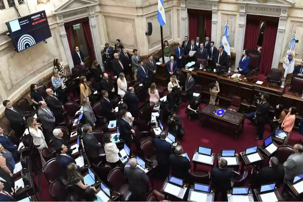 Overview of the Argentine Senate chamber with senators and staff standing during a session, surrounded by desks with computers and Argentine flags in the background Overview of the Argentine Senate chamber with senators and staff standing during a session, surrounded by desks with computers and Argentine flags in the background