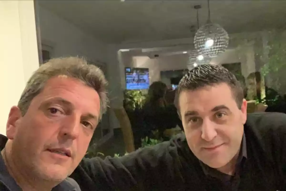 Dos hombres posan para una selfie en un ambiente interior iluminado con lámparas colgantes y un televisor encendido al fondo Dos hombres posan para una selfie en un ambiente interior iluminado con lámparas colgantes y un televisor encendido al fondo