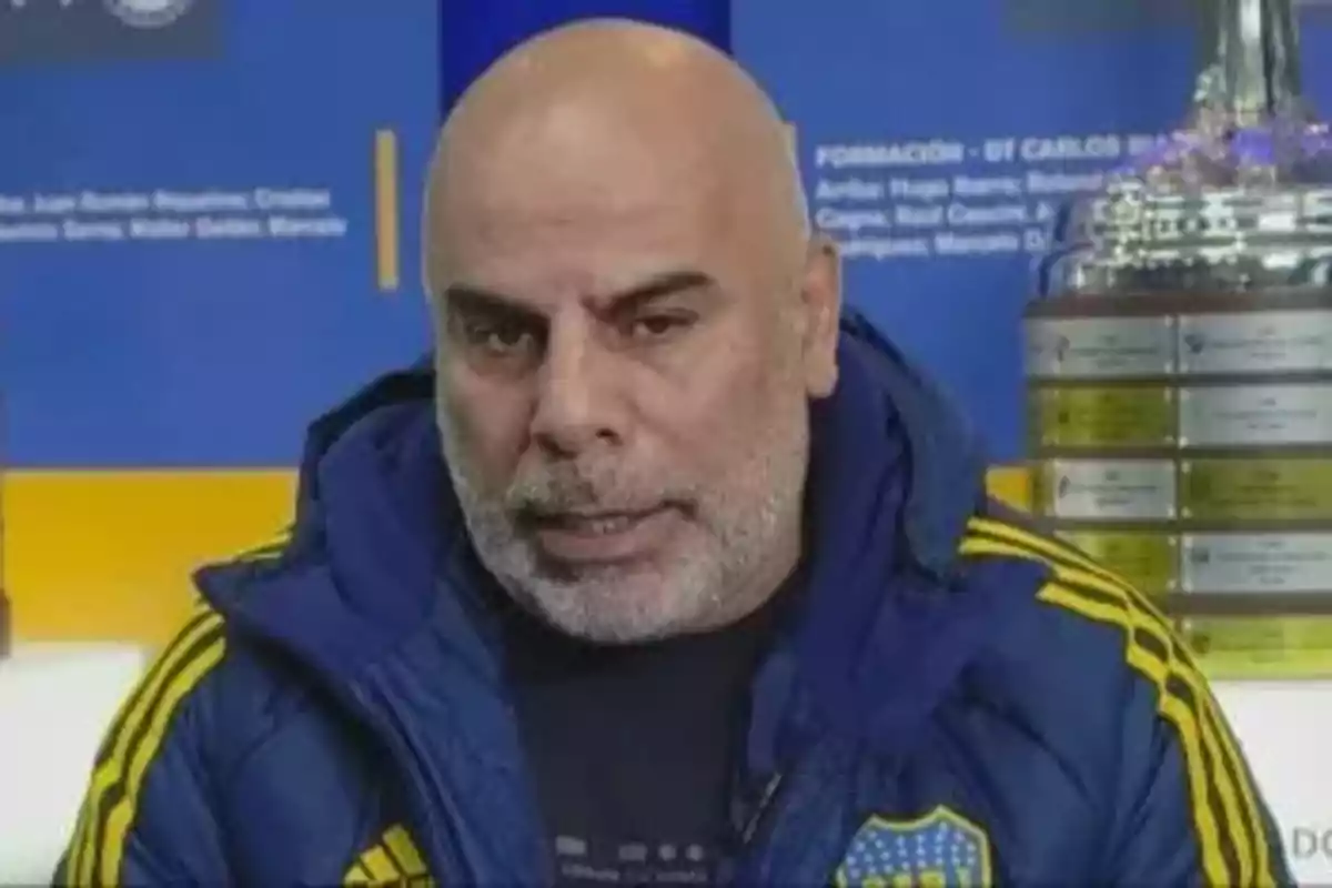 Hombre calvo con barba canosa vistiendo una campera azul de Boca Juniors con fondo azul y amarillo y un trofeo plateado a la derecha Hombre calvo con barba canosa vistiendo una campera azul de Boca Juniors con fondo azul y amarillo y un trofeo plateado a la derecha