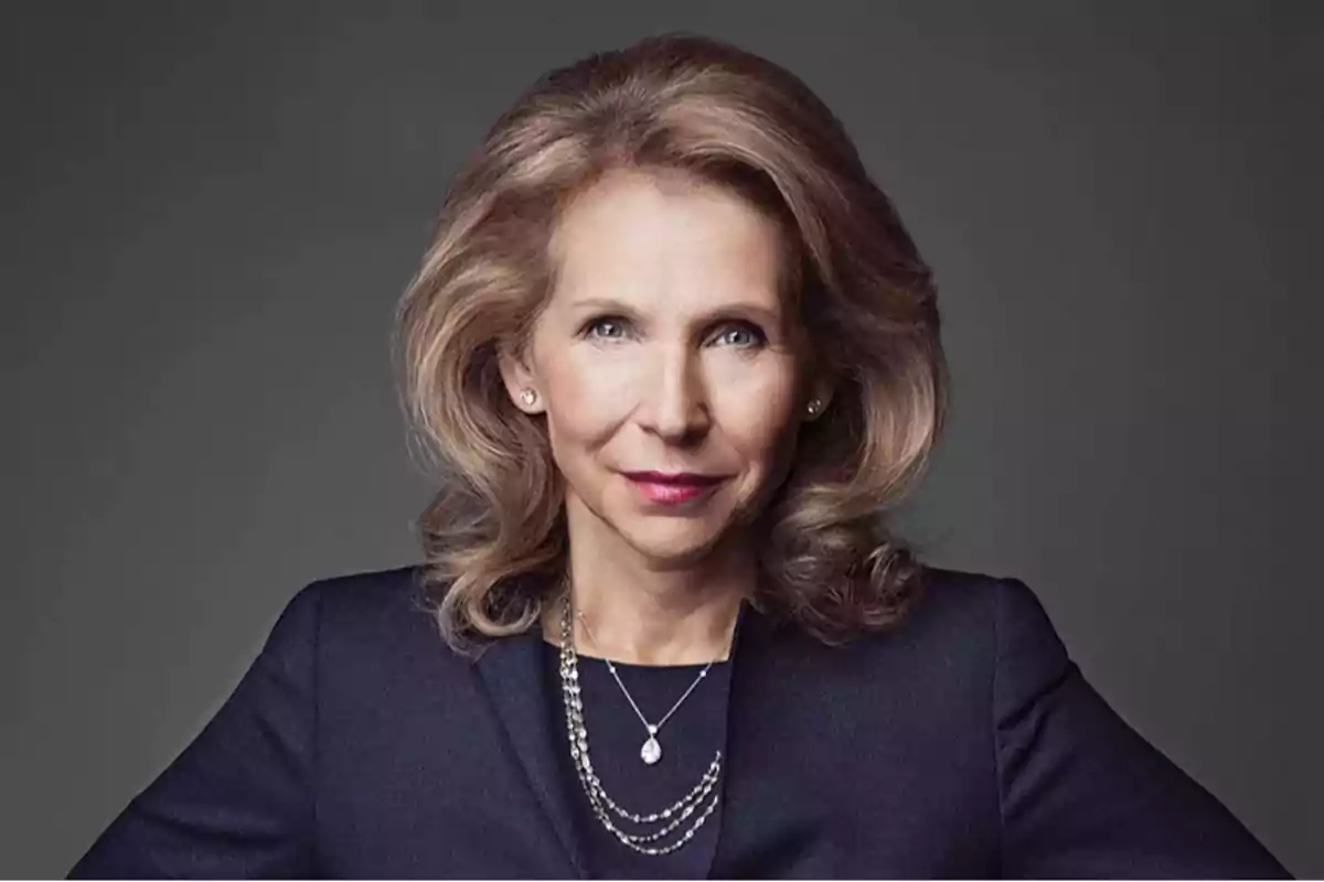 Blonde, wavy-haired woman wearing a dark blue blazer and a pearl necklace posing in front of a gray background Blonde, wavy-haired woman wearing a dark blue blazer and a pearl necklace posing in front of a gray background