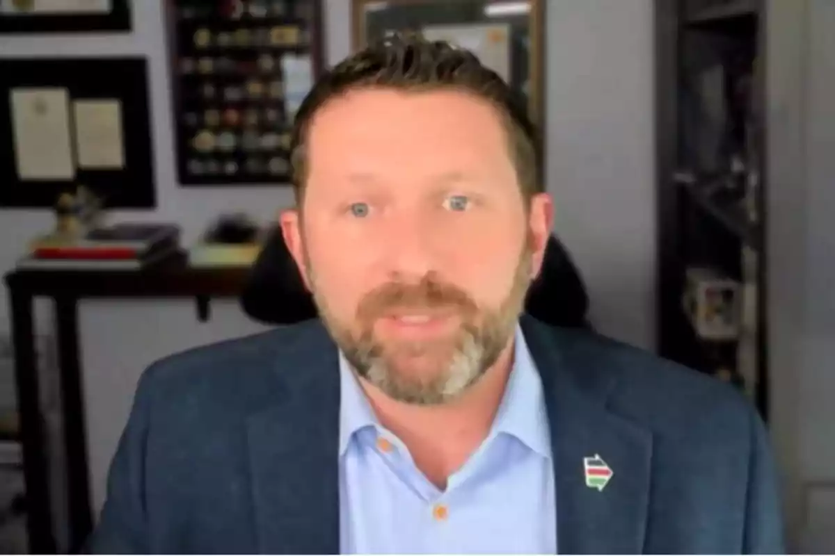 Hombre de barba y saco azul claro sentado en una oficina con estantes y cuadros al fondo Hombre de barba y saco azul claro sentado en una oficina con estantes y cuadros al fondo