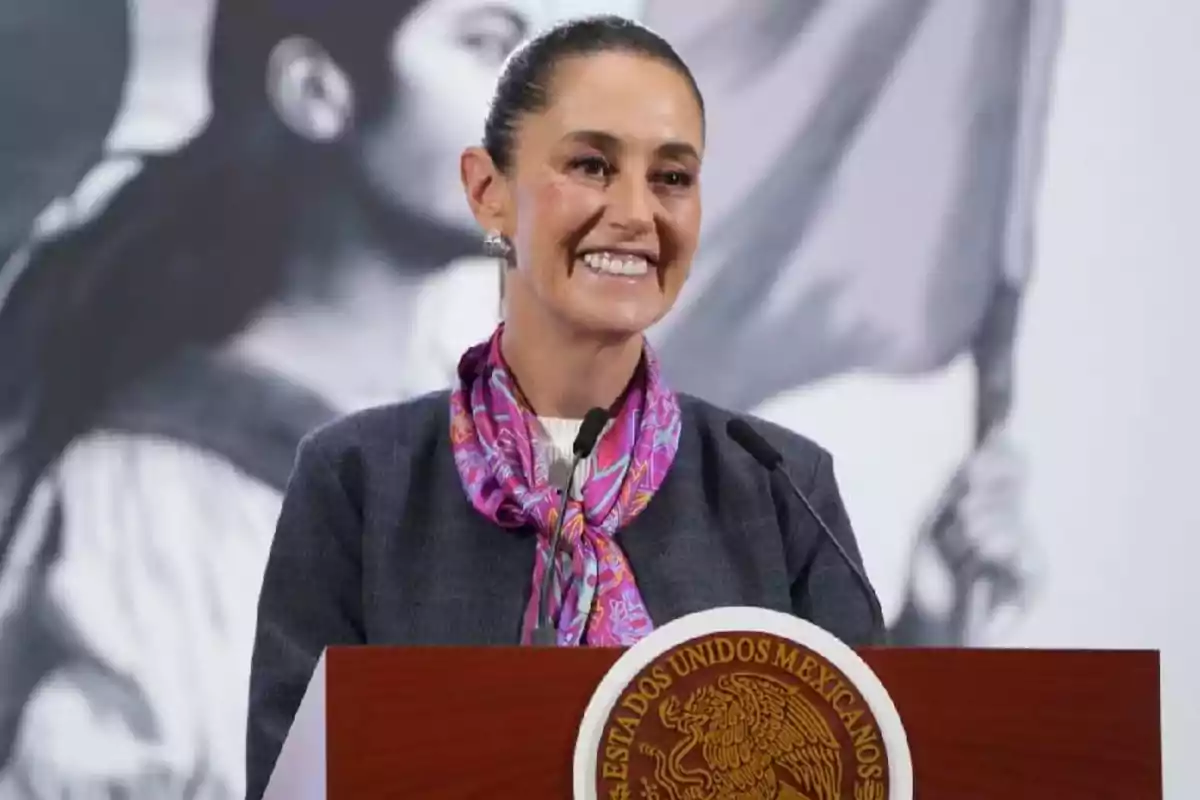 Mujer sonriente con saco oscuro y mascada rosa hablando en un podio con el escudo nacional de México al frente Mujer sonriente con saco oscuro y mascada rosa hablando en un podio con el escudo nacional de México al frente