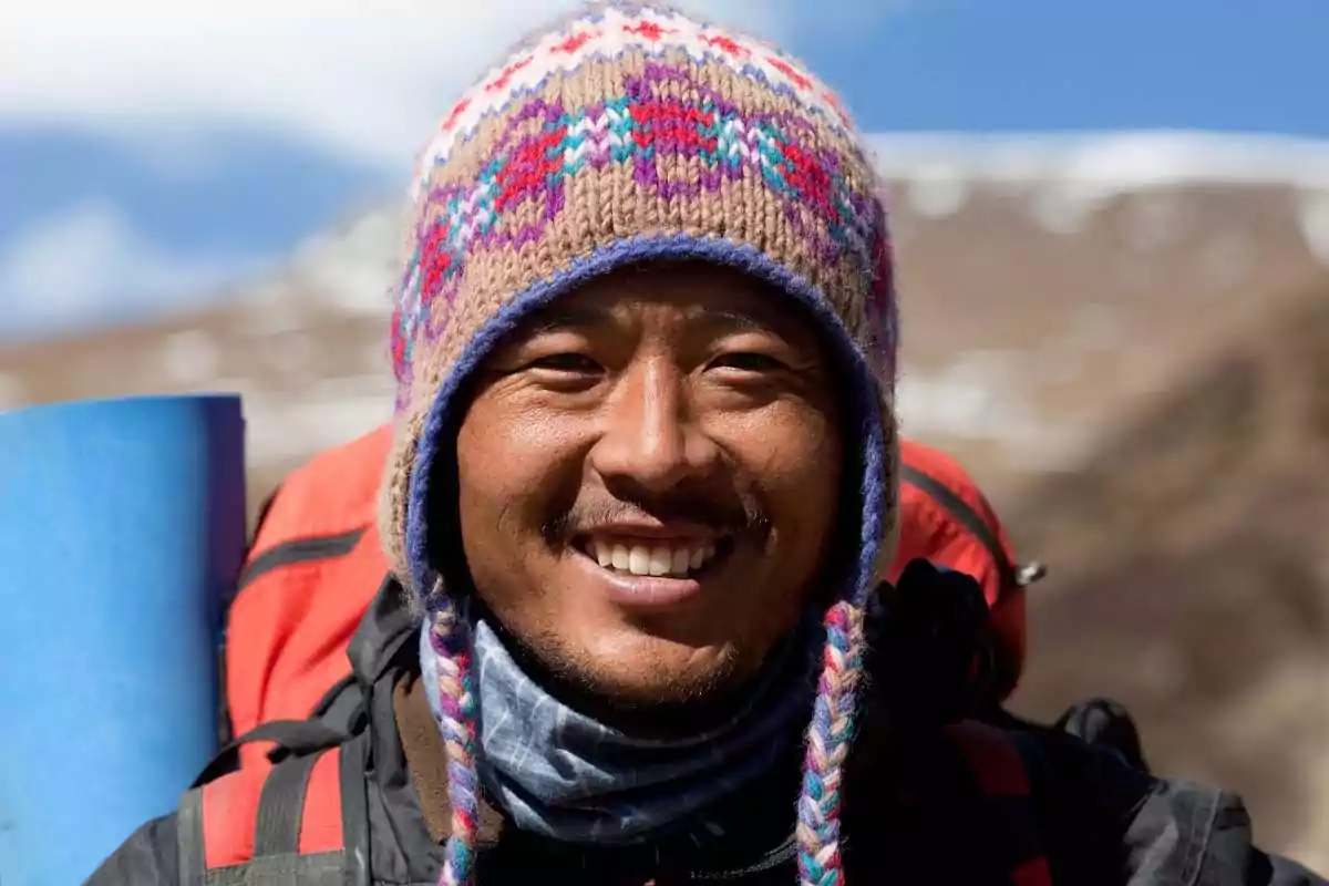Hombre sonriente con gorro de lana colorido y mochila en un entorno montañoso