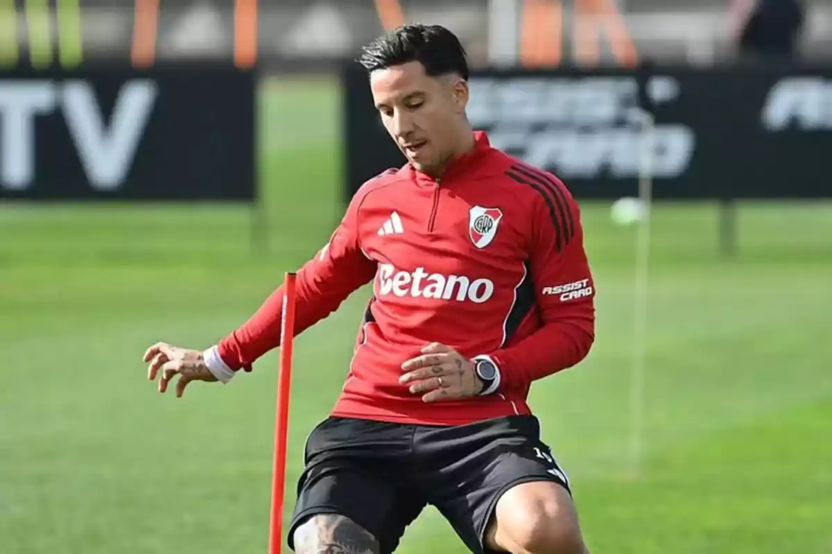 Futbolista de River Plate entrenando en el campo con ropa deportiva roja y negra esquivando un poste durante una práctica