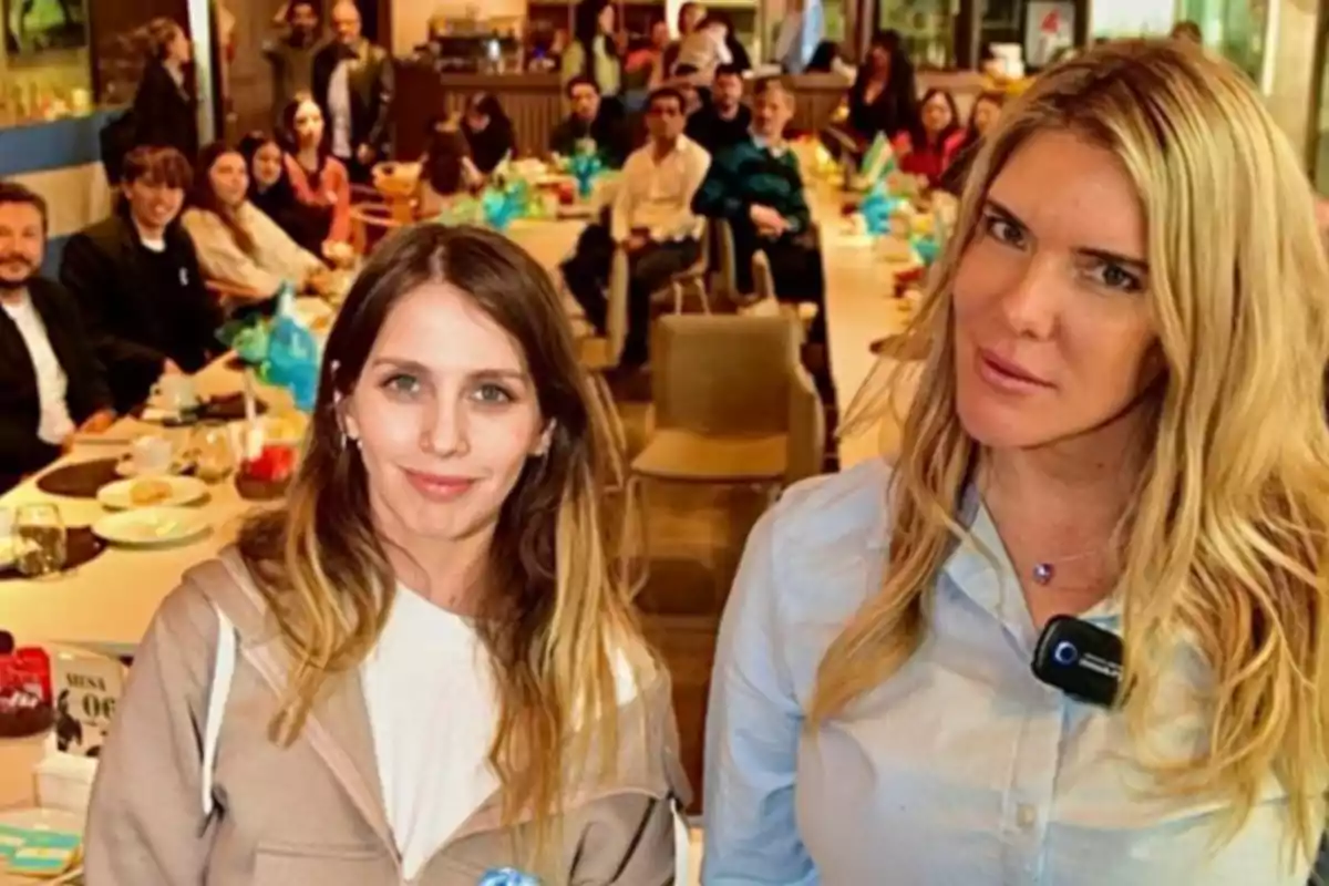 Dos mujeres posan al frente de una mesa larga en un restaurante rodeadas de varias personas sentadas y comida sobre la mesa