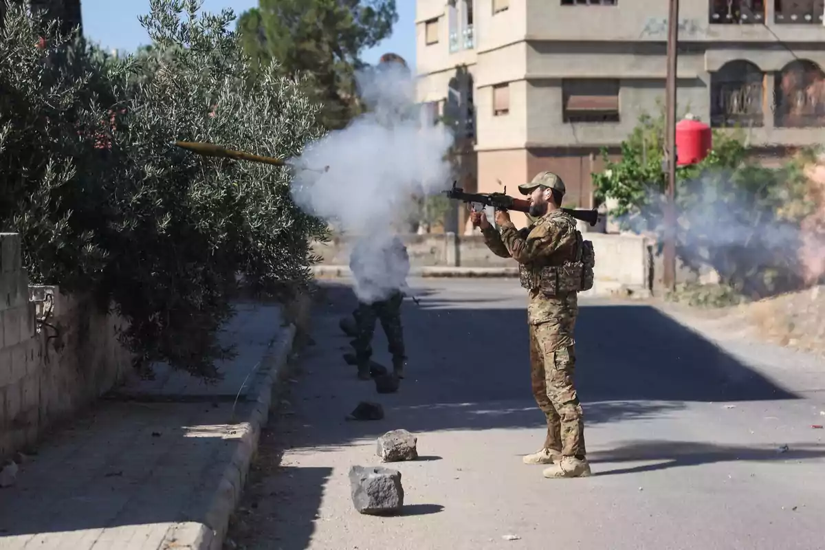 A soldier fires a rocket launcher on an urban street while another serviceman takes cover behind him, with smoke and rocks on the ground.