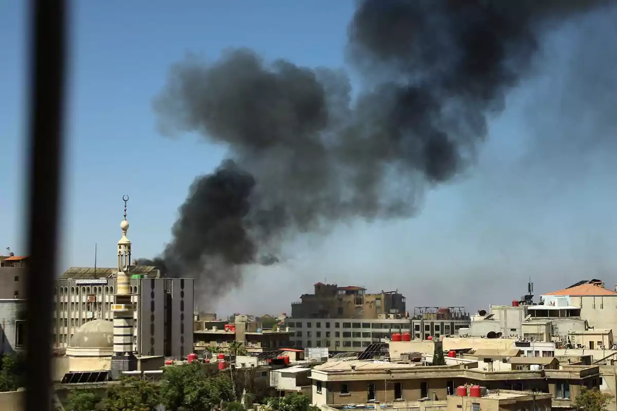 Black smoke rises over a city with buildings and a mosque under a clear sky.