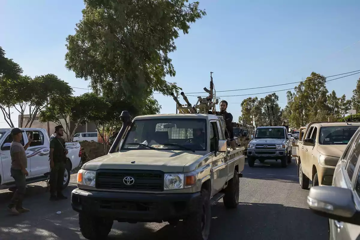 Several off-road vehicles drive down a street with armed men and trees in the background on a sunny day. Several off-road vehicles drive down a street with armed men and trees in the background on a sunny day.