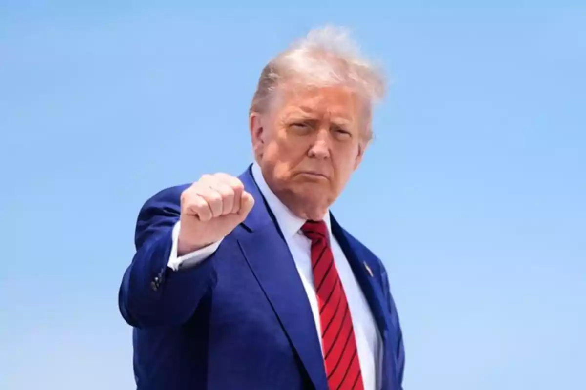 Blond-haired man in a blue suit and red tie raising his fist in front of a clear sky