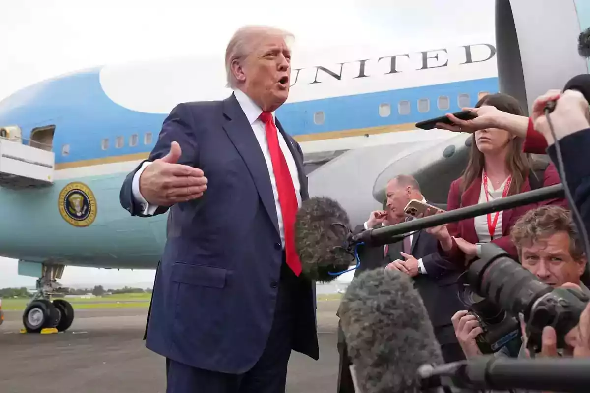 A man in a suit and red tie speaks in front of several microphones and cameras while a presidential plane is parked behind him. A man in a suit and red tie speaks in front of several microphones and cameras while a presidential plane is parked behind him.