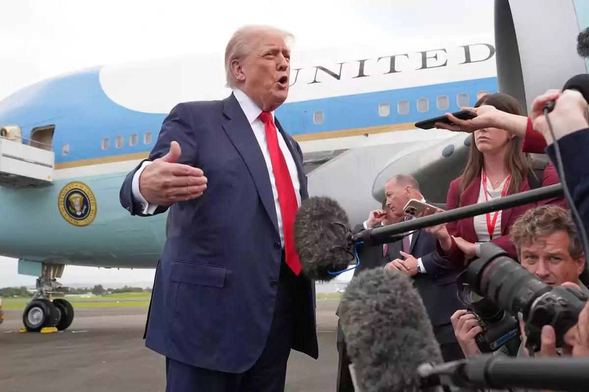 Man in a blue suit and red tie speaking in front of microphones and cameras with a presidential plane in the background