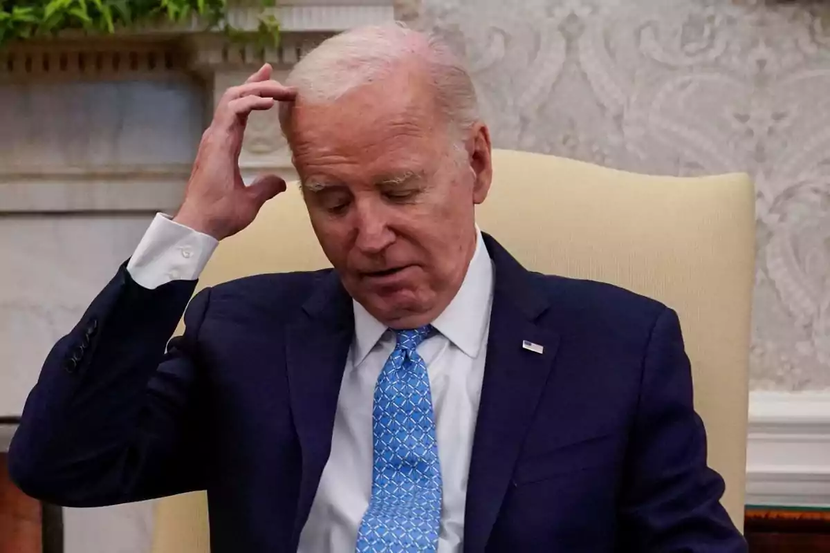 Older man with gray hair wearing a blue suit and light blue tie, sitting on a light-colored chair, touching his head with a thoughtful expression