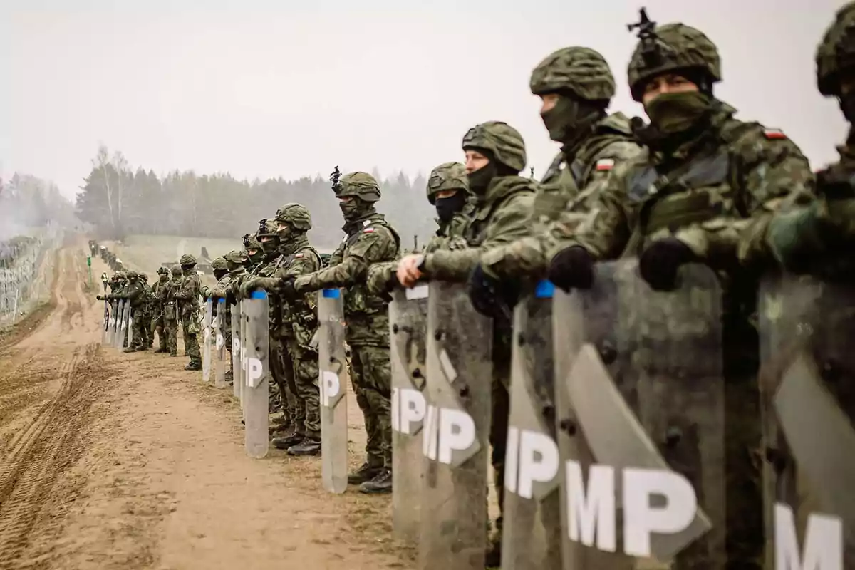 Soldiers with riot gear and shields lined up on a dirt road next to a fence in a wooded area. Soldiers with riot gear and shields lined up on a dirt road next to a fence in a wooded area.