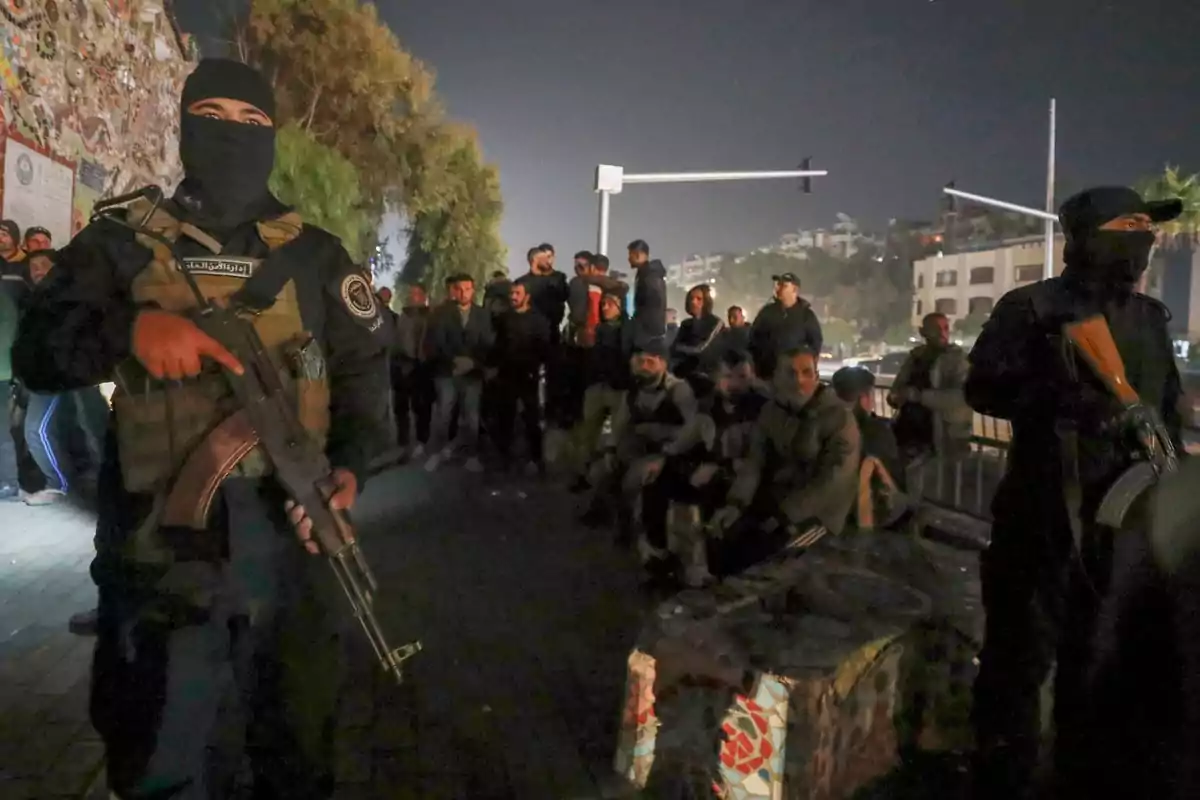 Policías armados con el rostro cubierto vigilan a un grupo de personas reunidas en la calle durante la noche Policías armados con el rostro cubierto vigilan a un grupo de personas reunidas en la calle durante la noche