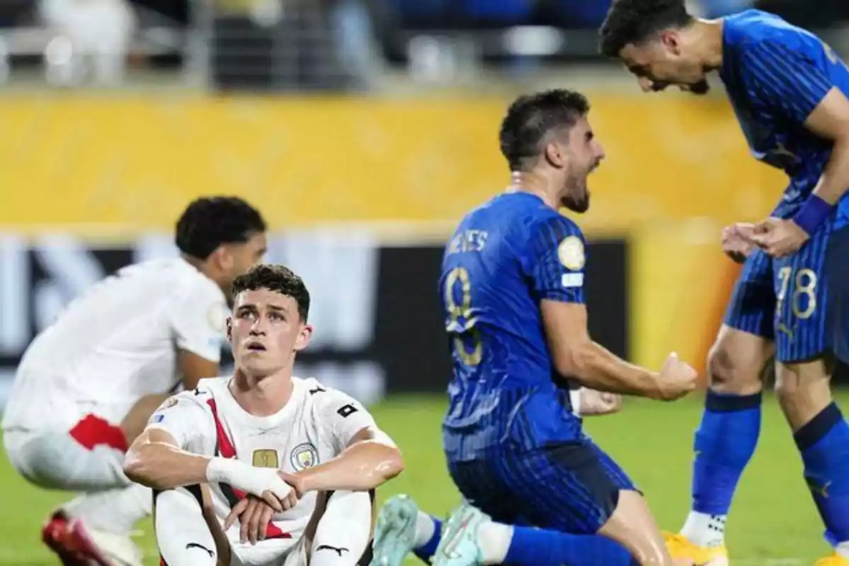 Jugadores de fútbol con uniformes blancos sentados en el césped mostrando desilusión mientras jugadores con uniforme azul celebran efusivamente en el fondo Jugadores de fútbol con uniformes blancos sentados en el césped mostrando desilusión mientras jugadores con uniforme azul celebran efusivamente en el fondo