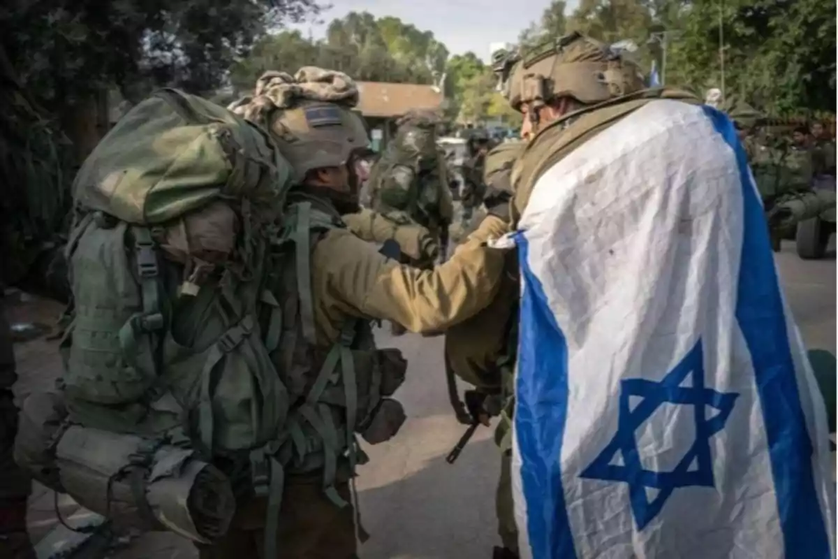 Soldiers with military gear walk together while one carries an Israeli flag over their shoulders.