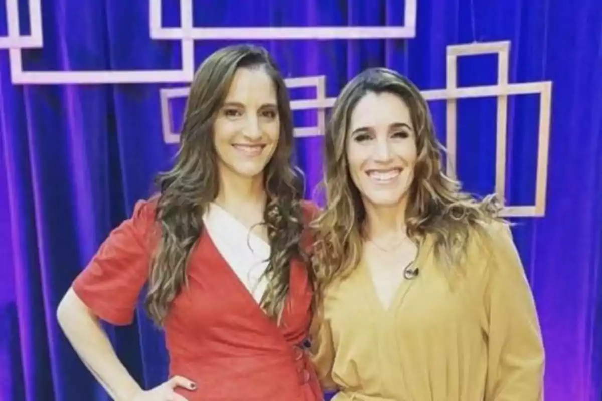 Dos mujeres sonríen frente a una cortina azul con figuras geométricas en el fondo una lleva vestido rojo y la otra vestido beige