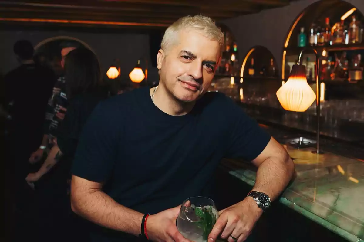 Short-haired, gray-haired man in a dark t-jersey smiles while holding a drink at the bar of a warmly lit venue with lamps and bottles in the background.
