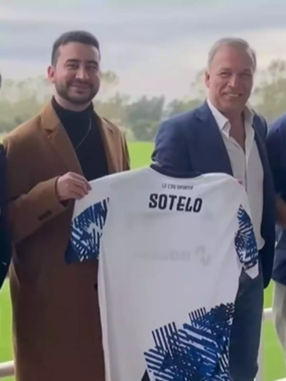 Dos hombres sonrientes sostienen una camiseta de fútbol blanca con detalles azules y el nombre Sotelo en la parte trasera Dos hombres sonrientes sostienen una camiseta de fútbol blanca con detalles azules y el nombre Sotelo en la parte trasera