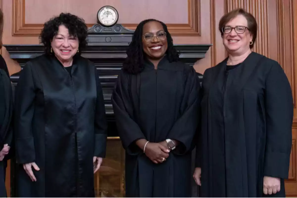Three women in black judges’ robes pose smiling in front of a wooden fireplace with an antique clock on top. Three women in black judges’ robes pose smiling in front of a wooden fireplace with an antique clock on top.