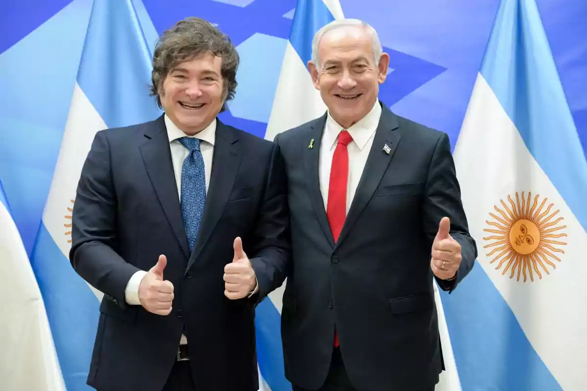 Dos hombres de traje sonríen y posan juntos haciendo el gesto de pulgar arriba frente a banderas de Argentina e Israel