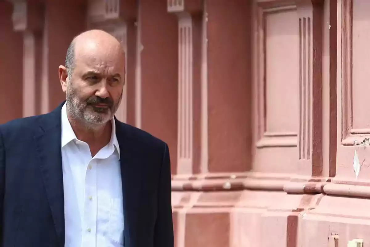 Hombre de mediana edad con barba y traje oscuro caminando junto a una pared rosada con detalles arquitectónicos