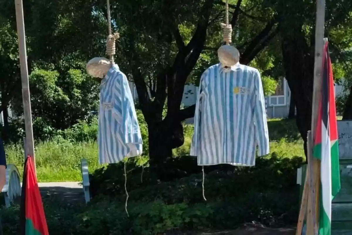 Dos figuras simulando personas ahorcadas con uniformes a rayas azules y blancas cuelgan de sogas en un entorno al aire libre rodeado de árboles y banderas.