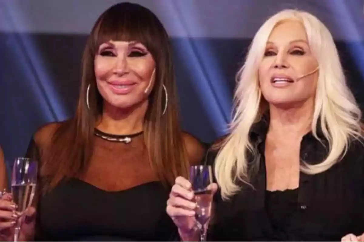 Two women smile and hold champagne glasses in a television studio setting.