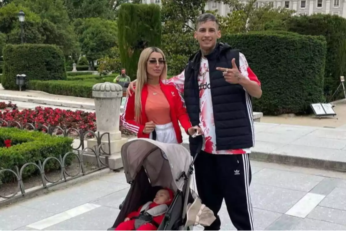 Una pareja joven posa en un parque junto a un cochecito donde duerme un bebé rodeados de jardines y arbustos bien cuidados