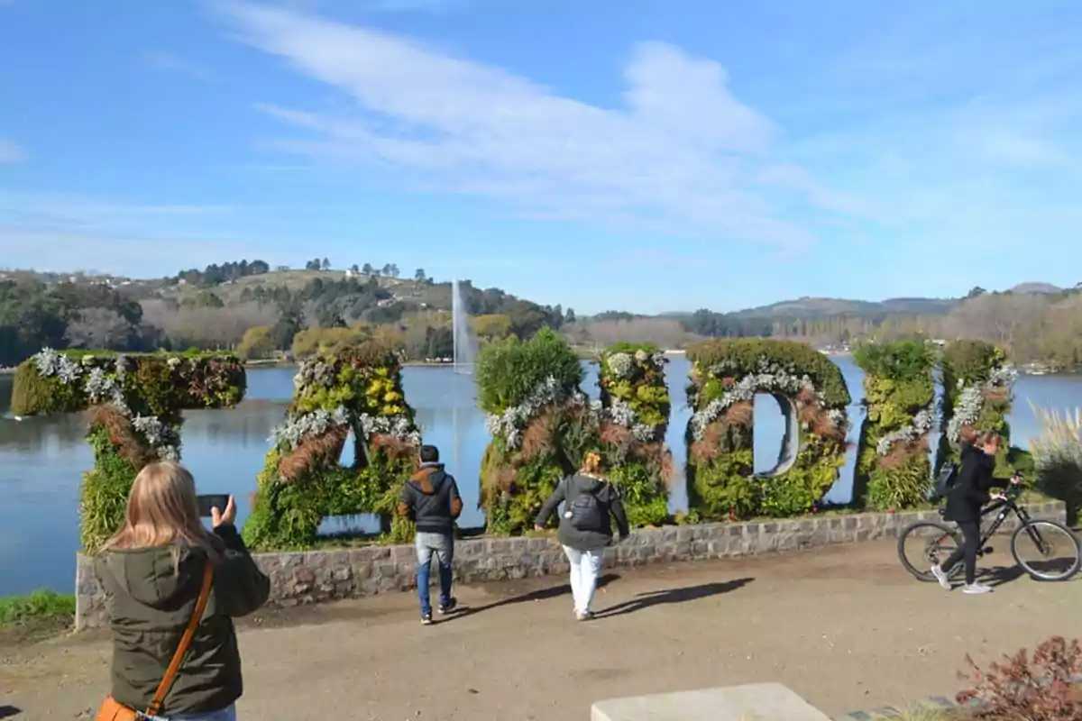 People walking and taking photos in front of a plant sign that forms the word Tandil, next to a lake with hills in the background. People walking and taking photos in front of a plant sign that forms the word Tandil, next to a lake with hills in the background.