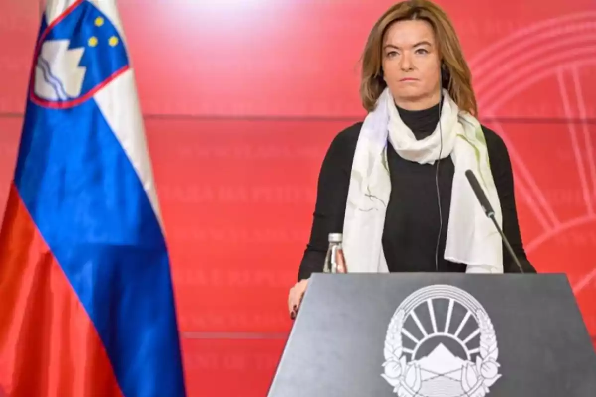 Brown-haired woman with a white scarf speaking at a podium in front of a Slovenian flag and a red background Brown-haired woman with a white scarf speaking at a podium in front of a Slovenian flag and a red background