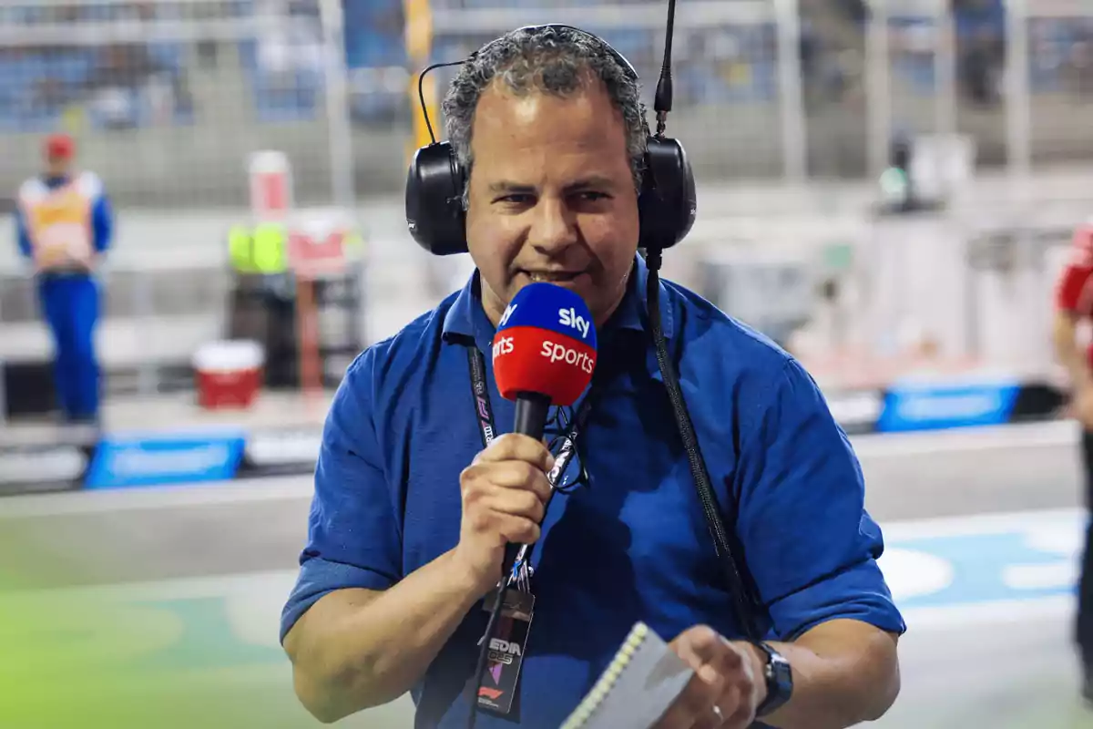 Hombre con auriculares grandes y camisa azul sosteniendo un micrófono de Sky Sports y una libreta en un entorno de pista deportiva Hombre con auriculares grandes y camisa azul sosteniendo un micrófono de Sky Sports y una libreta en un entorno de pista deportiva