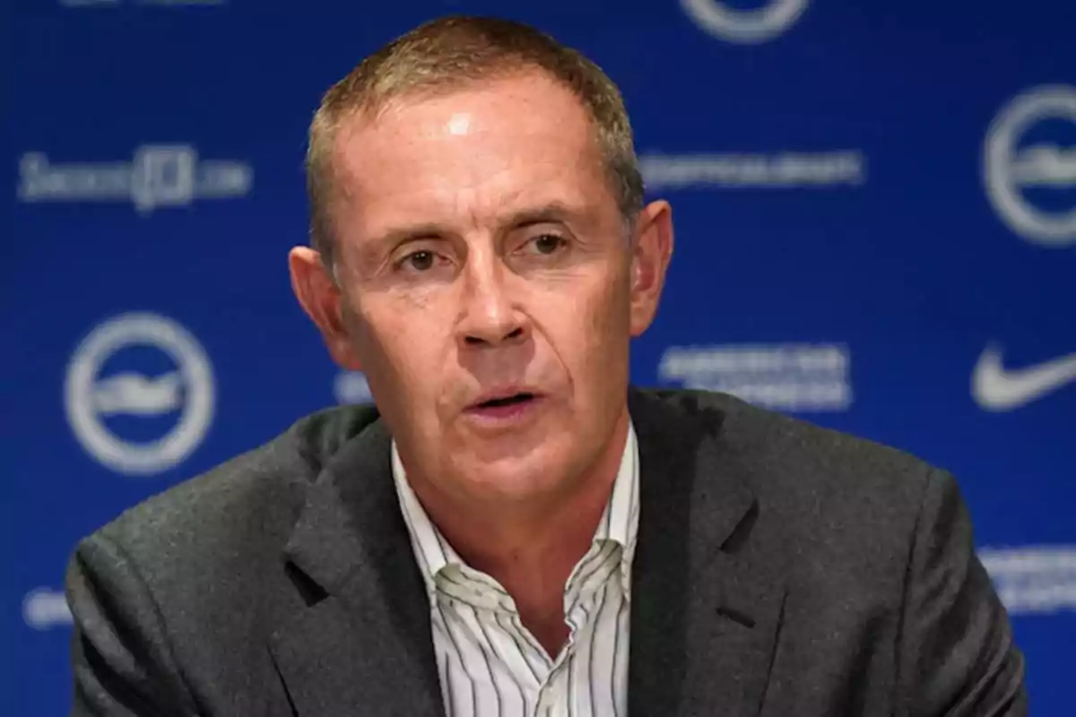 Hombre de mediana edad con cabello corto y canoso usando saco gris y camisa clara frente a un fondo azul con logotipos blancos Hombre de mediana edad con cabello corto y canoso usando saco gris y camisa clara frente a un fondo azul con logotipos blancos