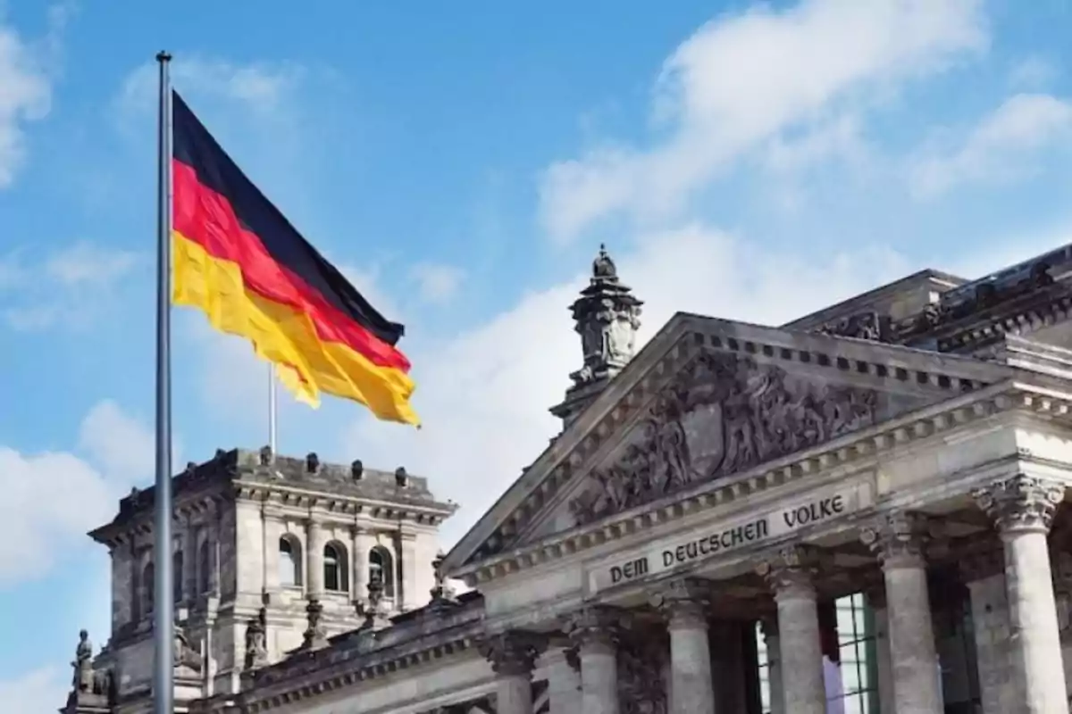 Bandera de Alemania ondeando frente al edificio del Reichstag en Berlín bajo un cielo azul con nubes. Bandera de Alemania ondeando frente al edificio del Reichstag en Berlín bajo un cielo azul con nubes.
